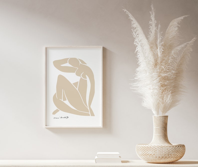 May include: A framed print of a beige abstract line drawing of a person sitting with their arm raised above their head. The print is on a white background and has a white frame. The print is hanging on a light beige wall.