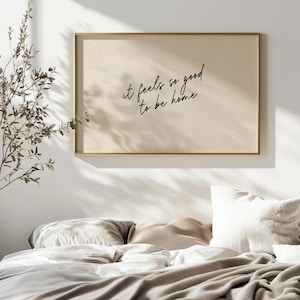 Peut inclure: Une affiche encadrée avec le texte "it feels so good to be home" dans un style manuscrit. L'affiche a un fond beige et un cadre doré. L'image est placée au-dessus d'un lit avec une literie blanche et beige.
