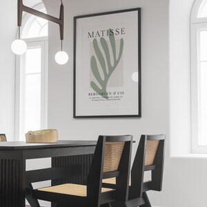 Op de afbeelding: Een zwart ingelijste print van een groen abstract kunstwerk van Matisse. De print hangt aan een witte muur boven een zwarte eettafel met twee zwarte stoelen met gevlochten rieten zittingen.