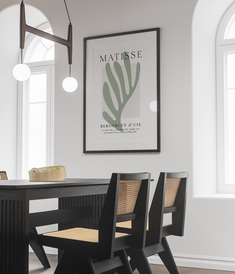 Op de afbeelding: Een zwart ingelijste print van een groen abstract kunstwerk van Matisse. De print hangt aan een witte muur boven een zwarte eettafel met twee zwarte stoelen met gevlochten rieten zittingen.