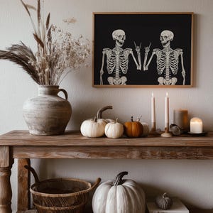 May include: A framed wall art print featuring two white skeletons on a black background, with both skeletons making a rock and roll hand gesture. The print is displayed in a wooden frame above a rustic wooden console table decorated with pumpkins.