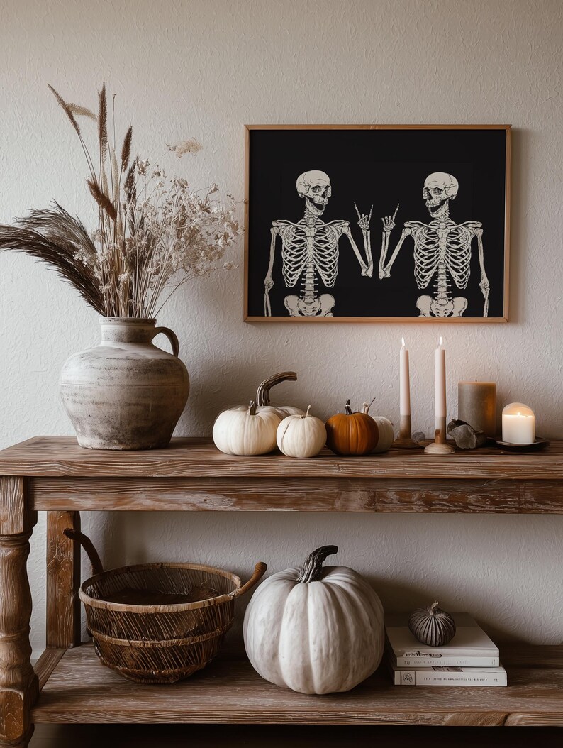 May include: A framed wall art print featuring two white skeletons on a black background, with both skeletons making a rock and roll hand gesture. The print is displayed in a wooden frame above a rustic wooden console table decorated with pumpkins.