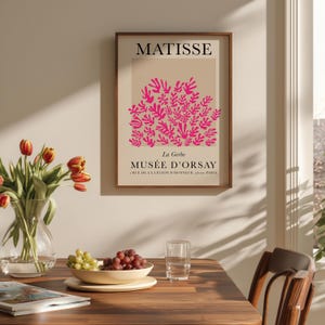 May include: A framed art print featuring the word "MATISSE" at the top, with a pink floral design and the text "MUS&Eacute;E D'ORSAY" below. The print is in a brown frame, displayed on a wall. A vase of tulips, a bowl of grapes, and a glass of water are on a wooden table.