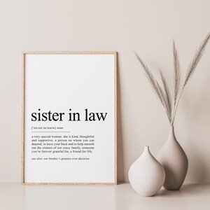 May include: A framed print with the words "sister in law" in a bold, black font. The definition of "sister in law" is below the title. Two neutral-toned vases and tall grass are to the right of the framed print.