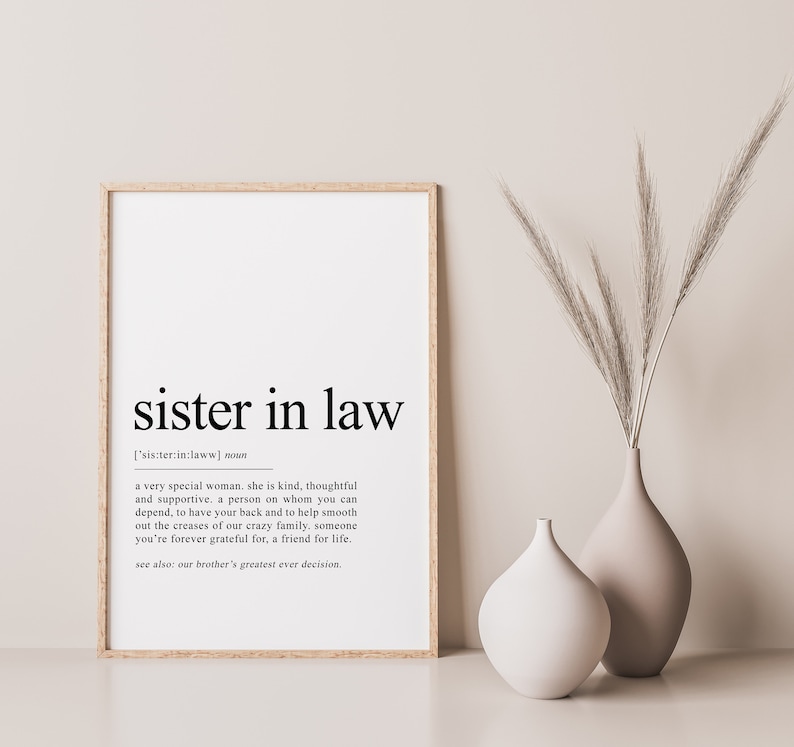 May include: A framed print with the words "sister in law" in a bold, black font. The definition of "sister in law" is below the title. Two neutral-toned vases and tall grass are to the right of the framed print.