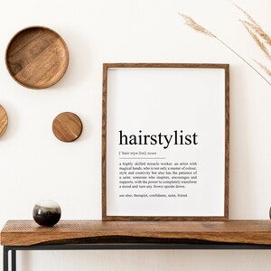 Puede incluir: Una impresi&oacute;n enmarcada con fondo blanco y texto negro que dice "hairstylist" y una definici&oacute;n de la palabra. La impresi&oacute;n est&aacute; colgada en una pared blanca con un estante de madera debajo. Hay art&iacute;culos decorativos de madera en el estante y un jarr&oacute;n con hierba seca.