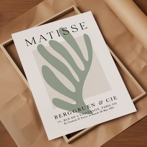 Op de afbeelding: Een minimalistische kunstprint met een groene abstracte vorm op een beige achtergrond. De print bevat de tekst "MATISSE" en "BERGGRUEN & CIE 70, RUE DE L'UNIVERSIT&Eacute;, PARIS-VII Du Vendredi 27 F&eacute;vrier au Samedi 28 Mars 1953."