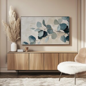 May include: A painting of a branch of eucalyptus leaves in shades of blue and green against a light beige background. The painting is framed and hanging on a wall above a wooden cabinet.