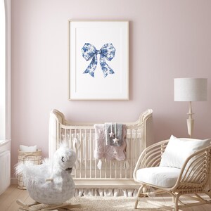 Puede incluir: Una cuna blanca con un caballo de balanc&iacute;n blanco y una silla de mimbre blanca con un coj&iacute;n blanco. La cuna tiene una colcha rosa y gris con estrellas. Hay una impresi&oacute;n enmarcada de un lazo azul y blanco en la pared.