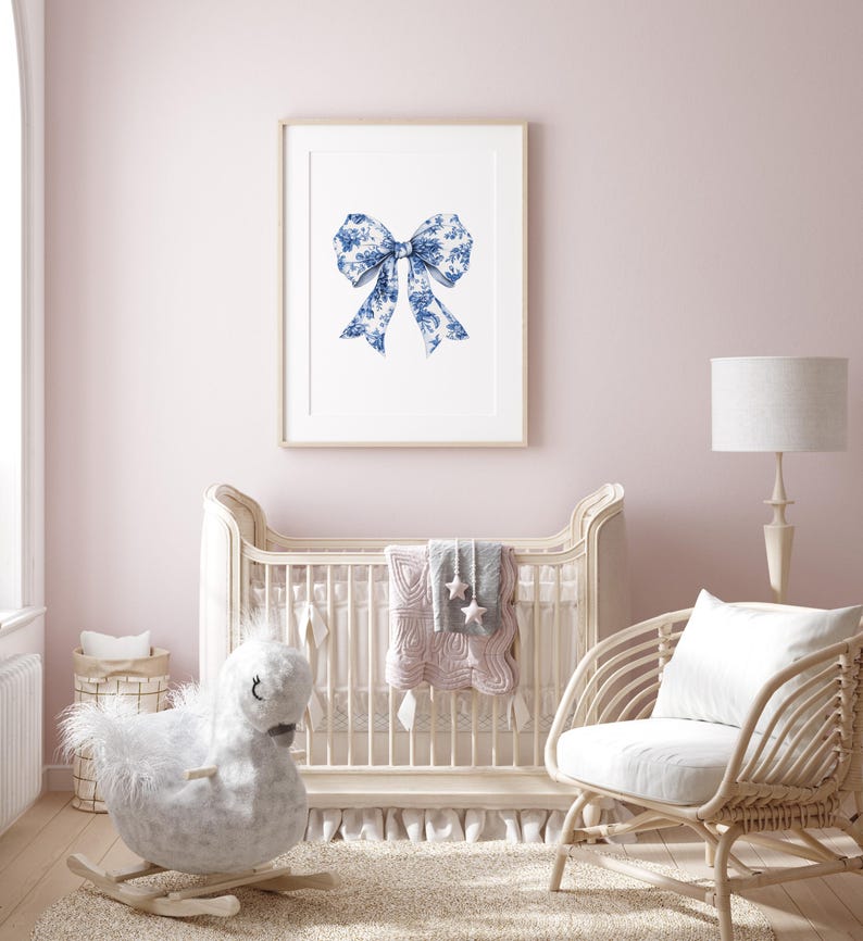 Puede incluir: Una cuna blanca con un caballo de balanc&iacute;n blanco y una silla de mimbre blanca con un coj&iacute;n blanco. La cuna tiene una colcha rosa y gris con estrellas. Hay una impresi&oacute;n enmarcada de un lazo azul y blanco en la pared.