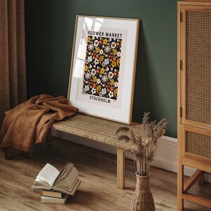 May include: A framed poster with a floral pattern in shades of pink, orange, yellow, and blue on a black background. The poster reads "FLOWER MARKET STOCKHOLM". The poster is hanging on a green wall above a wooden bench.