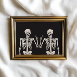 May include: A rectangular wall art print in a metallic gold frame featuring two off-white skeletons on a solid black background. Both skeletons are depicted from the waist up, each making a rock-and-roll hand gesture with one hand.