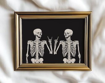 May include: A rectangular wall art print in a metallic gold frame featuring two off-white skeletons on a solid black background. Both skeletons are depicted from the waist up, each making a rock-and-roll hand gesture with one hand.