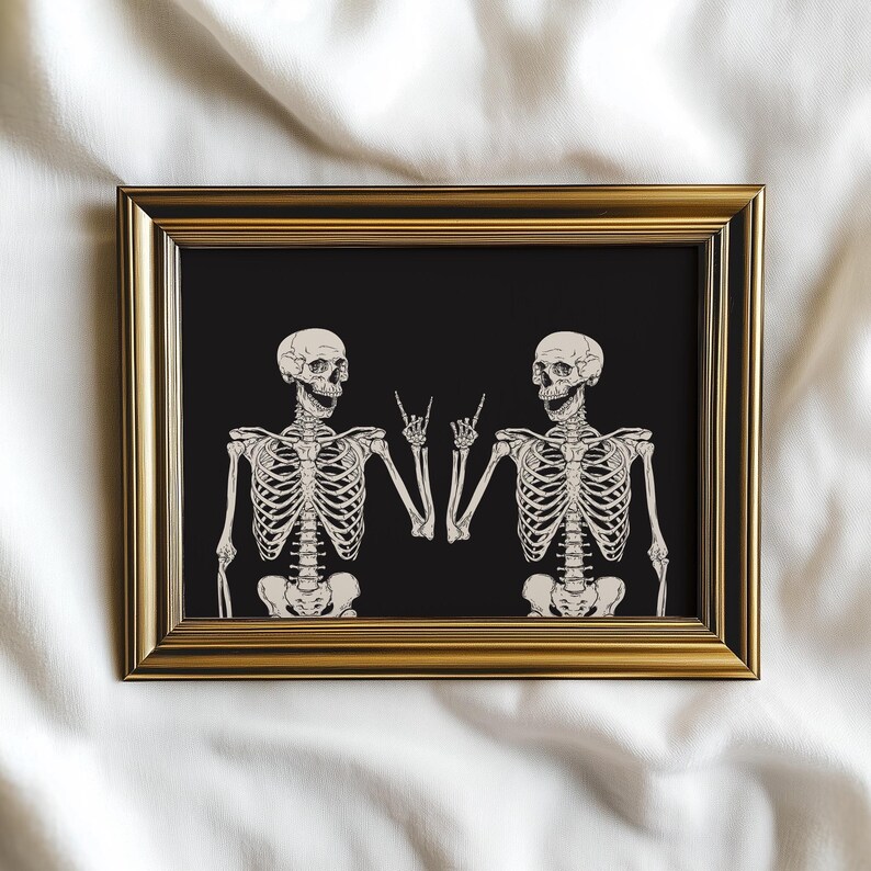 May include: A rectangular wall art print in a metallic gold frame featuring two off-white skeletons on a solid black background. Both skeletons are depicted from the waist up, each making a rock-and-roll hand gesture with one hand.