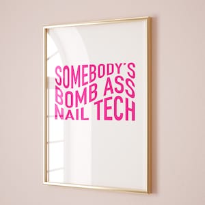 Puede incluir: Un póster enmarcado con el texto "SOMEBODY'S BOMB ASS NAIL TECH" en letras de color rosa brillante. El texto está dispuesto en un diseño lúdico y ligeramente inclinado sobre un fondo blanco. El marco es delgado, de color dorado.