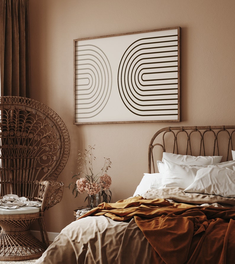 K&ouml;nnte beinhalten: Schlafzimmer-Interieur mit gerahmtem, abstraktem Kunstwerk. Das Kunstwerk hat einen braunen Holzrahmen und zeigt zwei halbkreisf&ouml;rmige Designs in Schwarz und Grau auf cremefarbenem Hintergrund. Ein Pfauenstuhl aus Korbgeflecht und ein Bett mit rostfarbener Decke sind ebenfalls zu sehen.