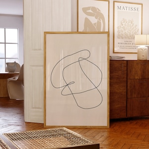 May include: A framed abstract line art print with a beige background and a dark blue line drawing. The print is in a wooden frame and is hanging on a wall with other framed prints.