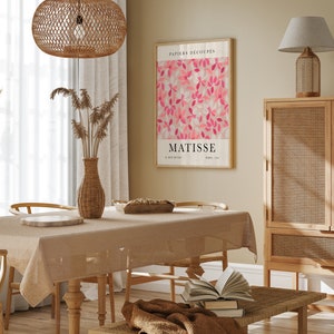 May include: A dining room with a wooden table and chairs, a wicker lamp hanging from the ceiling, a wicker cabinet, and a framed print of a pink floral design with the text "PAPIERS DECOUP&Eacute;S MATISSE 22 RUE DE BAC PARIS 1953".
