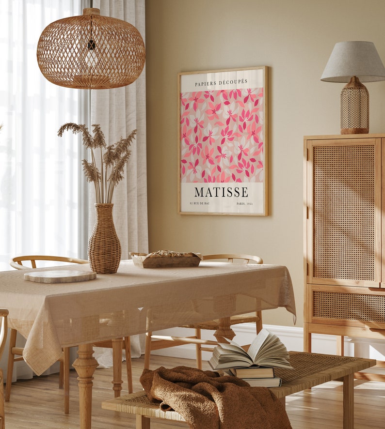May include: A dining room with a wooden table and chairs, a wicker lamp hanging from the ceiling, a wicker cabinet, and a framed print of a pink floral design with the text "PAPIERS DECOUP&Eacute;S MATISSE 22 RUE DE BAC PARIS 1953".