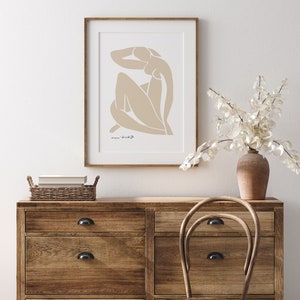 May include: A framed print of a beige abstract line drawing of a person with the text "marie bradburry" in the bottom left corner. The print is hanging on a white wall above a wooden dresser with drawers and a wicker basket on top. A wooden chair is in front of the dresser.