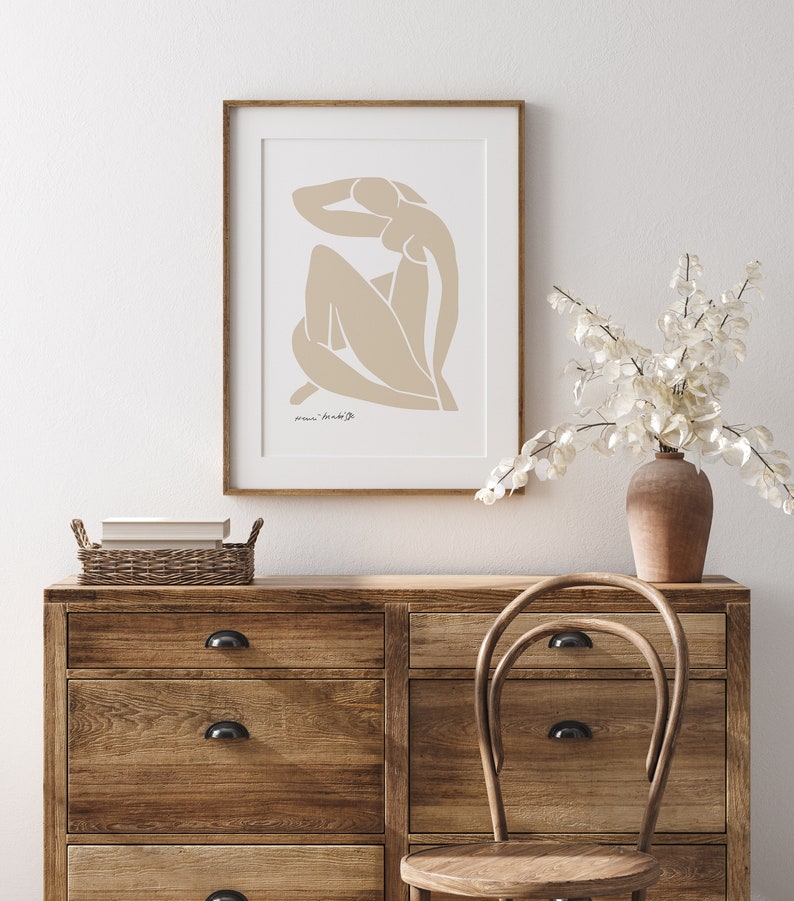 May include: A framed print of a beige abstract line drawing of a person with the text "marie bradburry" in the bottom left corner. The print is hanging on a white wall above a wooden dresser with drawers and a wicker basket on top. A wooden chair is in front of the dresser.
