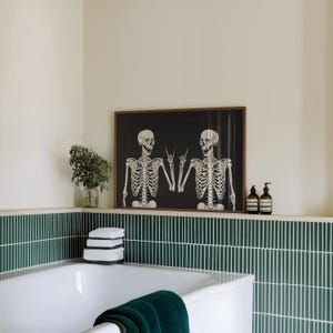 May include: A rectangular wall art print in a brown wooden frame featuring two white skeletons on a black background, both making a rock and roll hand gesture. The skeletons are positioned side by side, facing each other with open mouths.