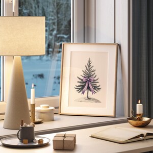 May include: A framed illustration of a small evergreen tree with a purple bow on a white background. The frame is on a wooden desk with a lamp, candles, and a book.