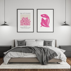 May include: A bedroom interior with a bed, two nightstands, a chair, and two framed prints on the wall. The prints are black and white with pink accents. The bed has a white duvet and a gray blanket. The nightstands are black and the chair is wooden.