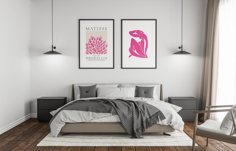 May include: A bedroom interior with a bed, two nightstands, a chair, and two framed prints on the wall. The prints are black and white with pink accents. The bed has a white duvet and a gray blanket. The nightstands are black and the chair is wooden.