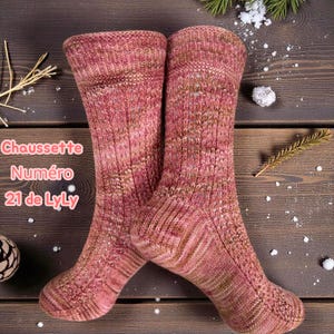 May include: A pair of hand-knitted, knee-high socks in shades of pink and brown. The socks feature a ribbed design and are displayed on a wooden surface with decorative elements. The text "Chaussette Numéro 21 de LyLy" is visible.