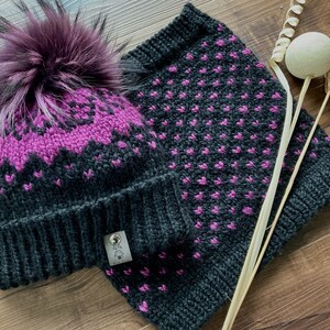 May include: A knitted hat and neck warmer set in charcoal gray and purple. The hat features a zig-zag pattern and a fluffy purple pom-pom. The neck warmer has a repeating pattern of small purple dots. The set is displayed on a wooden surface.