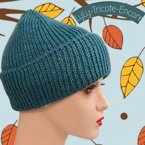 Peut inclure: Bonnet en tricot bleu sarcelle avec bord replié. Le chapeau est présenté sur une tête de mannequin avec du rouge à lèvres rouge. L'arrière-plan présente des feuilles d'automne et le texte "LyLy-Tricote-Encore".