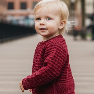 Peut inclure: Un cardigan en tricot bordeaux avec un motif texturé et une fermeture boutonnée. Le cardigan est porté par un jeune enfant aux cheveux blonds et un jean bleu. Le texte "Cardigan *Julien/Julienne*" est en haut de l'image.