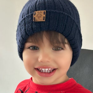 Może przedstawiać: Małe dziecko w niebieskim, dzianym berecie z brązową skórzaną naszywką z napisem "Lola" i "Little Dreamer". Dziecko uśmiecha się i ma na sobie czerwoną koszulkę z czarnym wzorem pajęczyny.