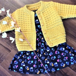 May include: A yellow knitted cardigan with button closures, laying on top of a black and white patterned dress.