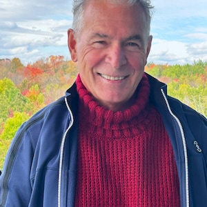 May include: A man with grey hair is smiling and wearing a red knitted turtleneck sweater under a blue zip-up jacket. He is standing in front of a background of autumn foliage.