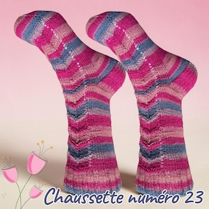 May include: A pair of hand-knitted socks featuring a variegated pattern of magenta, dusty rose, and slate blue horizontal stripes with a vertical chevron texture. The text "Chaussette numéro 23" is printed in dark blue at the bottom.