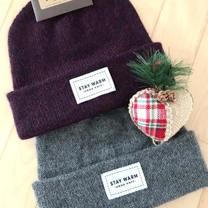 Op de afbeelding: Twee gebreide mutsen, één dieppaars en één grijs, worden getoond. Elke muts heeft een wit rechthoekig label met de tekst "STAY WARM - HAND KNIT-". Er is ook een klein decoratief hart met geruite stof en een dennentakje aanwezig.