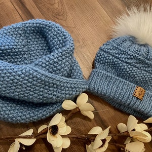 Könnte beinhalten: Eine blaue Strickmütze mit einem weißen Bommel und einem blauen Strick-Infinity-Schal. Die Mütze hat ein braunes Leder-Etikett mit dem Text "Made with Love" darauf.