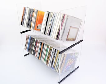 Puede incluir: Un estante de CD de acrílico transparente con dos estantes, lleno de varios CD. El estante tiene soportes negros y está ligeramente inclinado. Los CD tienen portadas coloridas. El fondo es blanco.