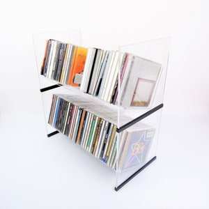 May include: A clear acrylic CD rack with two shelves, filled with various CDs. The rack has black supports and is angled slightly. The CDs have colorful album art. The background is white.