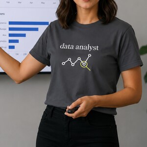 Peut inclure: Un t-shirt gris foncé à manches courtes avec le texte blanc "data analyst" au-dessus d'une icône de graphique linéaire blanc, incluant un cercle jaune soulignant l'un des points de données. Le t-shirt est porté avec un pantalon noir.