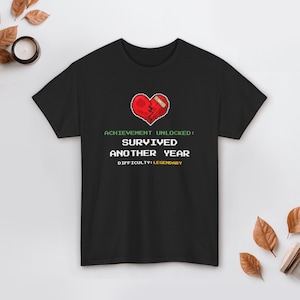 May include: Black t-shirt with a pixelated red heart graphic, a bandage, and the text "ACHIEVEMENT UNLOCKED: SURVIVED ANOTHER YEAR DIFFICULTY: LEGENDARY" in green and white. The shirt is a casual wear item.