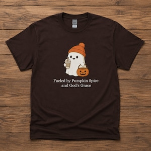 May include: A dark brown short-sleeved t-shirt featuring a graphic of a white ghost wearing an orange beanie, holding a coffee cup and a jack-o'-lantern bucket. Below the graphic, the text reads: Fueled by Pumpkin Spice and God's Grace.