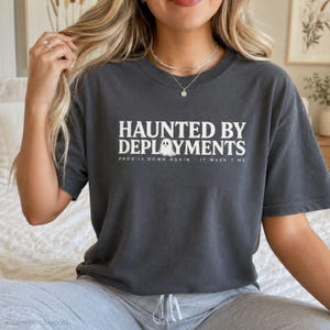 May include: A charcoal gray short-sleeve t-shirt featuring white text that reads HAUNTED BY DEPLOYMENTS with a small ghost graphic in the center and the smaller text PROD IS DOWN AGAIN IT WASN'T ME printed underneath the main graphic.