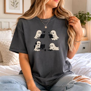 May include: A charcoal gray short-sleeve t-shirt featuring a graphic print of four white ghosts engaged in various activities, including using a laptop, holding a tablet, wearing headphones at a computer, and drawing on a digital tablet.