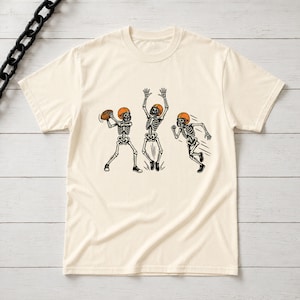 May include: A cream-colored t-shirt featuring a graphic of three skeletons wearing orange football helmets. One skeleton is throwing a football, one is jumping with arms raised, and the third is running. The skeletons are depicted in black ink.