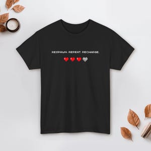 May include: Black t-shirt with the text "RESPAWN. REPEAT. RECHARGE." above four pixelated hearts, three red and one gray. The shirt is a classic crew neck style.