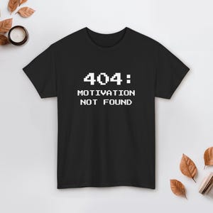 Könnte beinhalten: Schwarzes T-Shirt mit dem weißen Text "404: MOTIVATION NOT FOUND" in Pixel-Schrift. Das T-Shirt hat einen Rundhalsausschnitt und eignet sich für den täglichen Gebrauch. Das Design ist schlicht und auffällig, ideal für Freizeitkleidung.