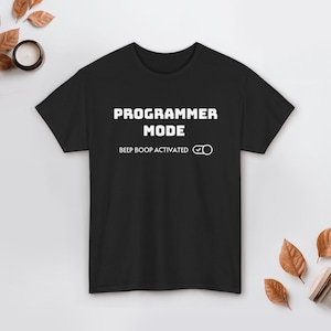 Pode incluir: Camiseta preta com a frase "PROGRAMMER MODE" em letras brancas. Abaixo, lê-se "BEEP BOOP ACTIVATED" com um símbolo de visto. A camiseta é de manga curta e gola redonda.